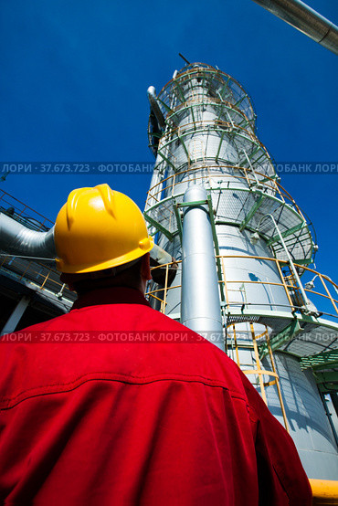 Refinery worker in red work wear and yellow hardhat on oil refinery and gas processing plant. Looking on Distillation column on blue sky background. (2012 год). Редакционное фото, фотограф Alexey Rezvykh / Фотобанк Лори