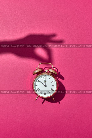 Retro alarm clock painted golden with hard shadows from woman’s hand holding it on a hot pink background, copy space.. Hand’s shadows holds retro golden painted alarmclock. Стоковое фото, агентство Ingram Publishing / Фотобанк Лори