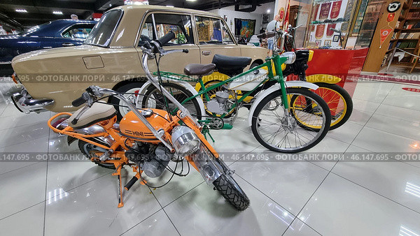 Retro vehicles from the Soviet era, displayed in the USSR Museum. The focus is on mopeds, motorcycles and a Soviet car.An orange mini-motorcycle with a low frame, compact design and a front headlight fairing.A green moped with a motor reminiscent of models like "Riga" or "Karpaty". It has a characteristic design of a bicycle with an engine.A yellow moped with chrome parts and a retro design.A Soviet car "Zhiguli" (VAZ-2101 or VAZ-2103) in beige with a chrome bumper.Retro motorcycles in the far corner with characteristic features of Soviet motor vehicles.An exhibition of items from the Soviet era, including awards, banners and rare books. (2023 год). Редакционное фото, фотограф Анна Кривицкая / Фотобанк Лори