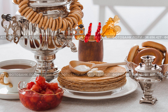 Russian traditions. Russian holiday Maslenitsa. Still life with a cup of tea, a stack of pancakes, sour cream, honey, cubes of butter, bagels, dried fruits, caramel cockerel and samovars. Стоковое фото, фотограф Photojuli / Фотобанк Лори