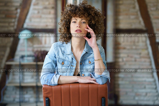Seductive young female, with opened mouth in black bra and denim jacket sitting on leather chair and touching face while looking at camera in loft style room. Sensual curly haired woman sitting on chair against brick wall. Стоковое фото, агентство Ingram Publishing / Фотобанк Лори
