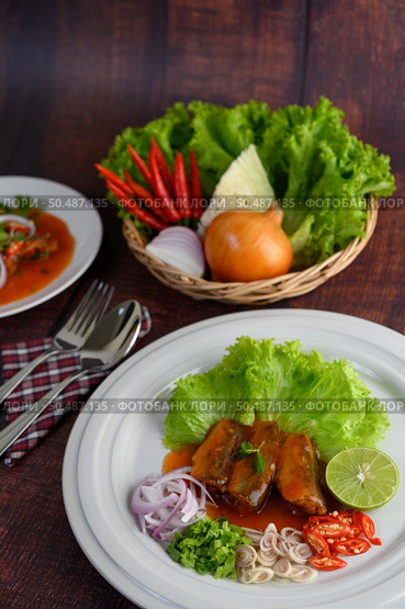 Selective focus Spicy salad of sardine with tomato sauce arranged nicely in white dish, ingredient herb for cooking in wicker basket, Stainless spoon and fork over napkin on wooden table, copy space. Стоковое фото, агентство Ingram Publishing / Фотобанк Лори