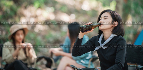 Selective focus young Asian pretty woman and her girlfriend sitting at front of tent, use mobile phone take photo during camping in forest with happiness together. Стоковое фото, агентство Ingram Publishing / Фотобанк Лори