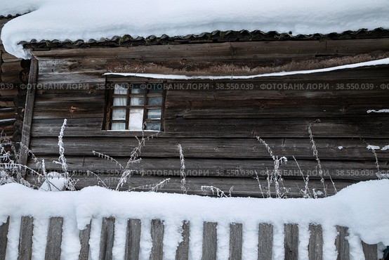 Село Домнино, Сусанинский район Костромской области. Зимний пейзаж. Маленькое окно на стене старого неокрашенного деревянного дома за утонувшим в негу забором (2022 год). Стоковое фото, фотограф Наталья Николаева / Фотобанк Лори