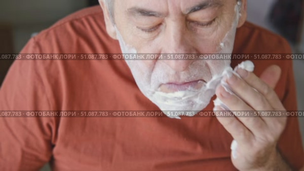 Senior Man shaving his face. Old male shaves beard and mustache with shaver. Elderly gray haired man looking at the mirror, applies shaving cream foam and using razor. Skin hygiene. Daily body care. Стоковое видео, видеограф Марина Демидюк / Фотобанк Лори