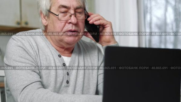 Serious senior man wearing glasses Talking mobile phone sitting at desk at home workplace with laptop. Retired male working chatting colleagues. Old worker talk on cellphone work on computer gadget. Стоковое видео, видеограф Марина Демидюк / Фотобанк Лори