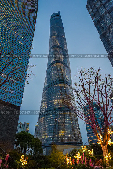 Shanghai, China – 1 April 2025: Twilight view of Shanghai Tower illuminated against dusk sky with glowing butterfly installations and floral decorations at its base. Редакционное фото, фотограф Татьяна Терехина / Фотобанк Лори