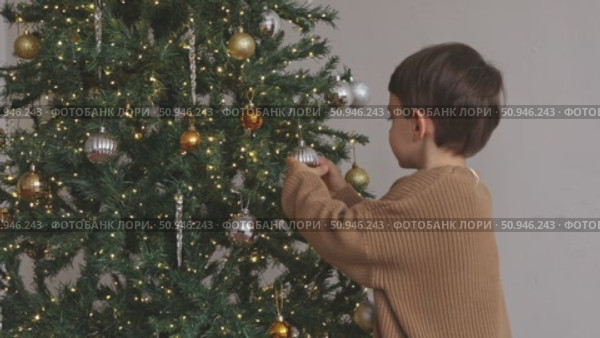 Side view of a cute little boy wearing a brown warm sweater, decorating a Christmas tree in the living room at home. Merry Christmas and happy New Year winter holidays. High-quality footage. Стоковое видео, видеограф Ирина Михайличенко / Фотобанк Лори