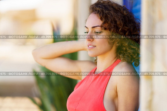 Side view of pensive young woman in sundress with hand touching back of head standing in street near wall with blue shutters and looking down against blurred urban background. Attractive young woman standing near blue shutters. Стоковое фото, агентство Ingram Publishing / Фотобанк Лори