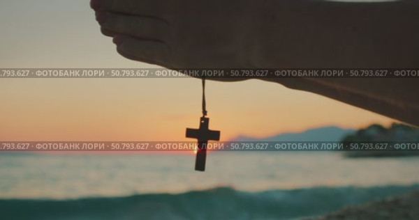Silhouette of praying hands holding a cross pendant in front of sea sunset with mountains in the distance. Стоковое видео, видеограф Igor Tichonow / Фотобанк Лори