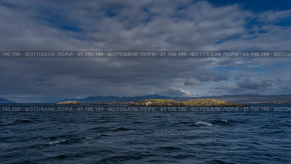 Small rocky islets with sparse stunted vegetation are visible in the ocean. Andes mountain range against the sky and clouds. Waves, foam on blue water. Argentina. Beagle Channel. (2023 год). Стоковое фото, фотограф Вера Ивановна Кубарева / Фотобанк Лори
