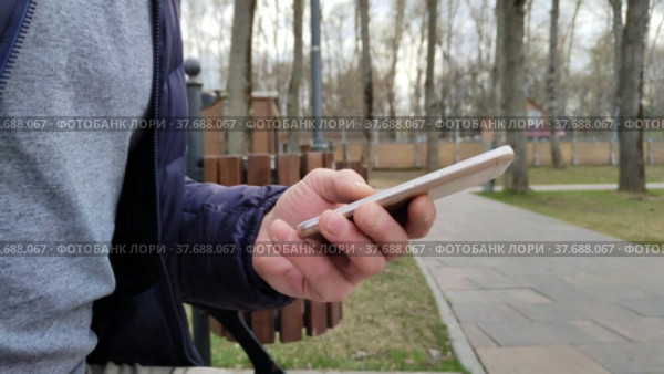 Смартфон в мужской руке крупным планом. Молодой мужчина, сидящий на скамейке в парке, пишущий текстовое сообщение или загружающий приложение для мобильного телефона. Рука набирает СМС. Стоковое видео, видеограф Ирина / Фотобанк Лори