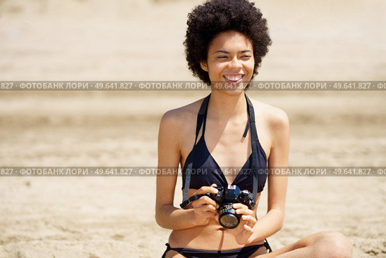 Smiling African American female tourist in stylish bikini with professional photo camera looking away while sitting on sandy shore in tropical resort. Cheerful black photographer in swimwear sitting on beach. Стоковое фото, агентство Ingram Publishing / Фотобанк Лори