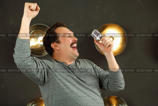 Smiling man with mustache sings with microphone in studio with lamps on wall. Стоковое фото, фотограф Losevsky Pavel / Фотобанк Лори