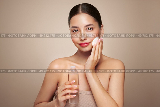 Smiling woman holds bottle with cosmetic tonic. Photo of attractive woman with perfect makeup on beige background. Beauty and skin care concept. Removing makeup. Стоковое фото, агентство Ingram Publishing / Фотобанк Лори