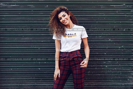 Smiling young arabic woman with black curly hairstyle. Arab girl in casual clothes in the street. Happy female wearing white t-shirt and checked pants against urban blinds. Стоковое фото, агентство Ingram Publishing / Фотобанк Лори