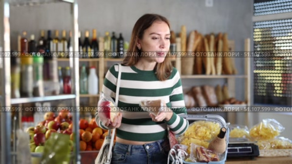 Smiling young girl with shopping basket trying to make choice holding two sorts of cheese in grocery store with varied stock. Стоковое видео, видеограф Яков Филимонов / Фотобанк Лори