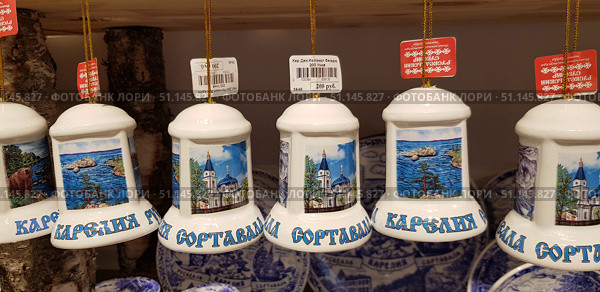 Souvenir ceramic bells with images of Karelia's landmarks, in particular the city of Sortavala. Each bell is decorated with colorful pictures - an Orthodox church with blue domes, landscapes with lakes, rocks and pine trees, as well as images of wildlife, such as a bear. The inscription "Karelia Sortavala" is written at the bottom of the bells. The souvenirs are suspended on golden cords, and price tags are visible at the top. The background is wooden elements and other goods with national motifs. The photo conveys the atmosphere of tourist souvenirs, folk art and regional identity. (2019 год). Редакционное фото, фотограф Анна Кривицкая / Фотобанк Лори