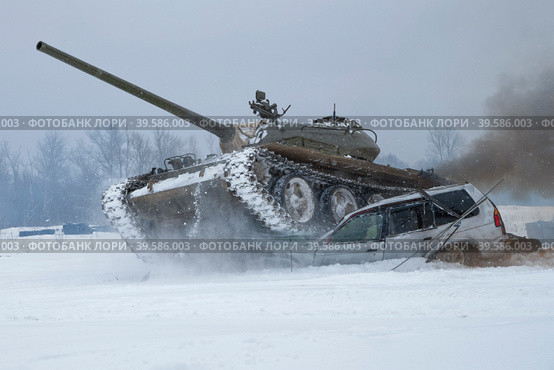 Советский танк Т-54 давит старый легковой автомобиль в заснеженном поле. Танковое шоу в военно-историческом парке "Стальной десант" (2023 год). Редакционное фото, фотограф Виктор Карасев / Фотобанк Лори