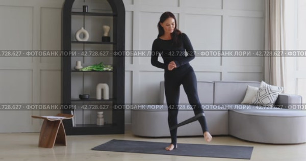 Спортивная женщина брюнетка занимается спортом дома, выполняет упражнения с фитнес резинкой, приседает чтобы укрепить мышцы ног, тренируется в черном спортивном комбинезоне на коврике, концепция здорового образа жизни. Стоковое видео, видеограф Сергей Чайко / Фотобанк Лори
