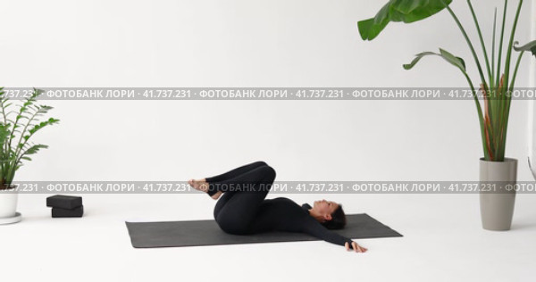 Спортивная женщина практикующая йогу, выполняет упражнение Павана Муктасана, позу освобождения ветра, укрепляет мышцы живота, тренируется в спортивном комбинезоне лежа на коврике в студии. Стоковое видео, видеограф Сергей Чайко / Фотобанк Лори