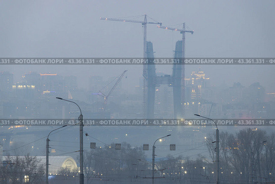 Строительство Центрального моста в Новосибирске. Башенные краны около строящейся высокой опоры вантового моста через реку Обь. Город Новосибирск, Новосибирская область, Сибирь, Россия. Вечерние сумерки и дымка. (2023 год). Редакционное фото, фотограф Андрей Степанов / Фотобанк Лори
