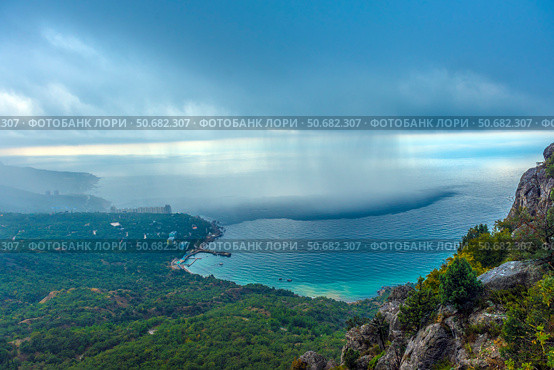 Stunning view of the Crimean coast from above. In the foreground you can see a rocky area covered with dense green vegetation, smoothly descending to the shore of the Black Sea. In the background are small buildings and residential complexes located along the coastal zone.The most impressive detail of the photo is the rain front hanging over the sea. The boundary between the clear and rainy zone is clearly visible, where streams of rain descend from the clouds and create the effect of a blurred horizon. This spectacle emphasizes the unique microclimate of Crimea, where mountains, sea and clouds form unpredictable weather phenomena. (2021 год). Стоковое фото, фотограф Анна Кривицкая / Фотобанк Лори