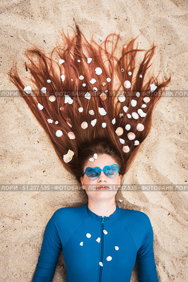 Stylish summer portrait of young girl lying on sand with seashells in her long hair, wearing heart-shaped sunglasses. Concept of creative holiday vibes, beach lifestyle, and youth fashion. Стоковое фото, фотограф Енваев Андрей / Фотобанк Лори