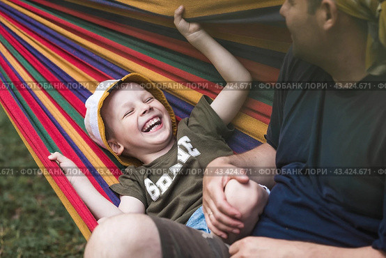 Summer holidays for children in the fresh air. A cute little boy with his father is having fun on a hammock in the backyard. A child and a dad in a hammock. Стоковое фото, фотограф Photojuli / Фотобанк Лори