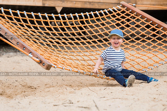 Summer lifestyle. A cute little boy is having fun playing in a hammock on the beach on a modern playground. The concept of family entertainment and leisure. Стоковое фото, фотограф Photojuli / Фотобанк Лори