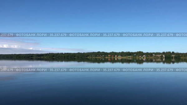 Surface of large forest lake with blue sky reflecting in calm water. Trees and village are on shore in countryside. Karelia, Russia. Стоковое видео, видеограф Кекяляйнен Андрей / Фотобанк Лори