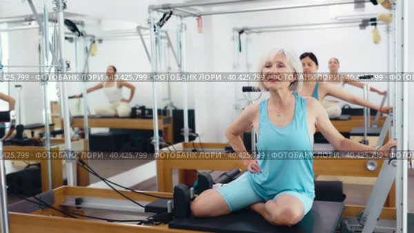 Svelte elderly woman performing set of pilates exercises on reformer during group workout. Active lifestyle and wellness concept of seniors. Стоковое видео, видеограф Яков Филимонов / Фотобанк Лори