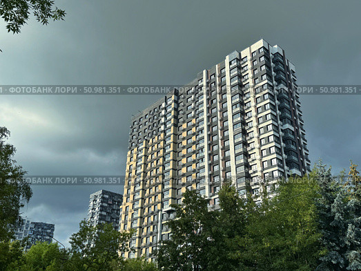 Tall apartment buildings against a dramatic, cloudy sky seen through green trees. Concept of: Urban living. Стоковое фото, фотограф Ольга Петрова-Апостолова / Фотобанк Лори