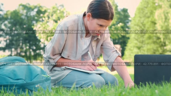 Teenage schoolgirl studying reading her books, tablet and notebook, sitting outdoors. Back to school. Student girl lying on the green grass using laptop in the college yard or park. Distance learning. Стоковое видео, видеограф Марина Демидюк / Фотобанк Лори