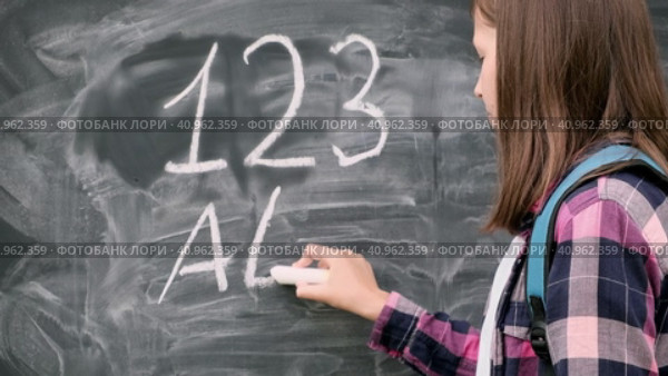 Teenage student girl in shirt with blue backpack writing with chalk numbers and letters on black chalkboard background. Education, back to school and people concept. Joyful Teen girl near blackboard. Стоковое видео, видеограф Марина Демидюк / Фотобанк Лори