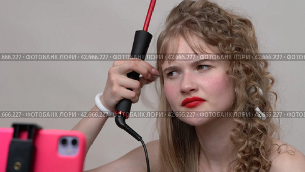 The blogger is using a curling wand to style hair by manually wrapping long hair around the wand. Woman sharing the process with us through a live stream on phone (2024 год). Редакционное видео, видеограф А. А. Пирагис / Фотобанк Лори