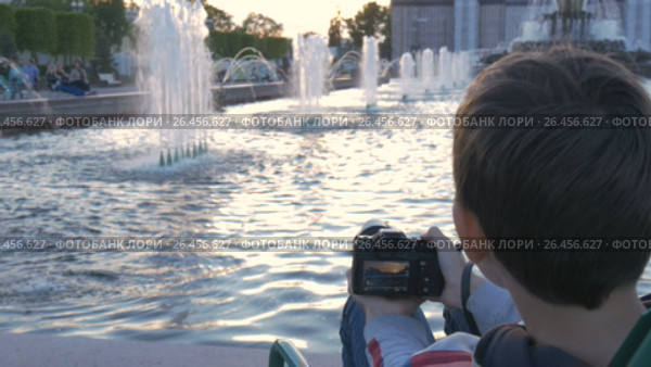 The boy is a teenager, sits in a beautiful spot by the fountain, and photographs water splashes. Beautiful landscape at sunset. A good place for rest and relaxation. Стоковое видео, видеограф Игорь Усачев / Фотобанк Лори