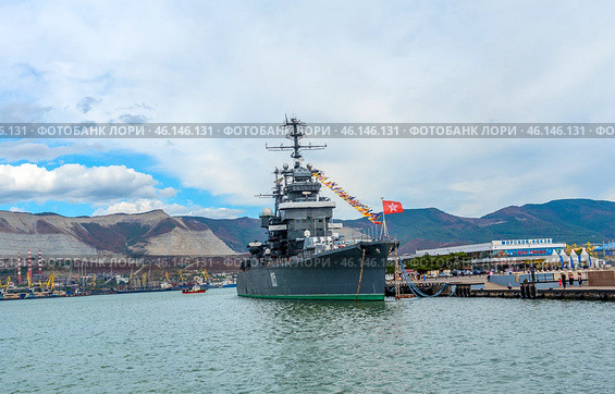 The cruiser Mikhail Kutuzov, permanently moored at the Novorossiysk embankment. This is one of the main museum ships of Russia, which is part of the Central Naval Museum.The cruiser Mikhail Kutuzov is a massive warship with the number "105" on board.It is decorated with signal flags, creating a festive atmosphere.In the background, the port of Novorossiysk, the mountains of the Markotkhsky Range and the berthing facilities are visible.The red naval flag with a white star flutters at the stern.To the right of the ship is the marine terminal, where people and city infrastructure are visible.In the distance, a small tug or boat moves along the water. (2023 год). Редакционное фото, фотограф Анна Кривицкая / Фотобанк Лори