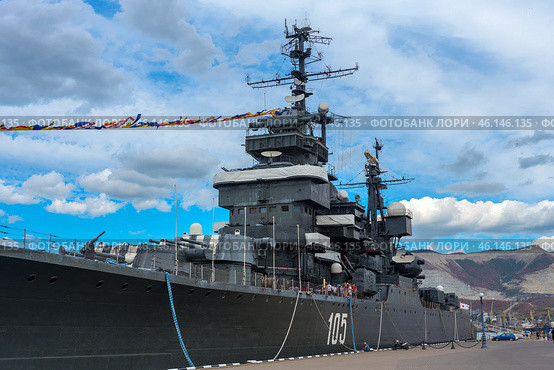 The cruiser "Mikhail Kutuzov", which is a museum ship in the city of Novorossiysk. This Project 68-bis warship was part of the USSR Navy for a long time, and then was turned into a museum, becoming one of the main attractions of the city.The side of the ship with the number "105", which indicates its affiliation with the Navy.You can see the flag garland - a ship decoration used during celebrations.Antennas and radar installations on the masts of the cruiser.A group of tourists on board, inspecting the ship.Mooring ropes secured to the pier.The protective paint of the hull is a characteristic dark gray color. (2023 год). Редакционное фото, фотограф Анна Кривицкая / Фотобанк Лори