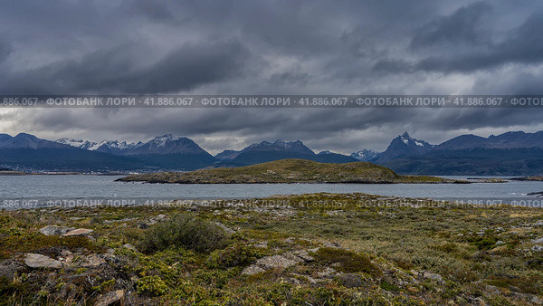 The landscape of southern Patagonia. On the islets in the Beagle Channel, sparse low-growing vegetation grows on rocky soil. The snow-capped mountain range of the Andes Martial against a cloudy sky. Стоковое фото, фотограф Вера Ивановна Кубарева / Фотобанк Лори