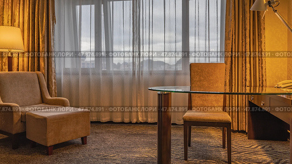 The living room, shades of beige. An upholstered chair at a glass table. An armchair with a banquette. Carpet on the floor. The lamp are lit. The mountains are visible through the window tulle. Стоковое фото, фотограф Вера Ивановна Кубарева / Фотобанк Лори