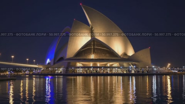 The modern architecture of Dubai Opera is reflecting in the water at night, with a bridge and city lights in the background, in Dubai, United Arab Emirates. Стоковое AI-видео, аниматор Данил Руденко / Фотобанк Лори