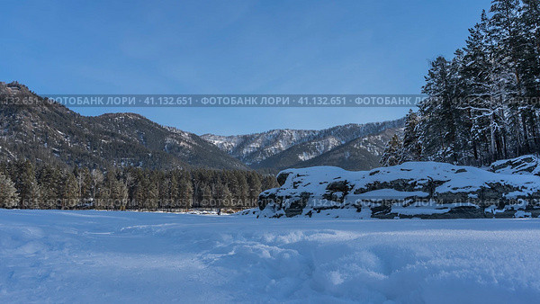 The path is trampled through snowdrifts. Fluffy hoarfrost sparkles. A layer of snow on picturesque boulders. Coniferous trees of the taiga and mountains against the blue sky. Copy space. Altai. Стоковое фото, фотограф Вера Ивановна Кубарева / Фотобанк Лори