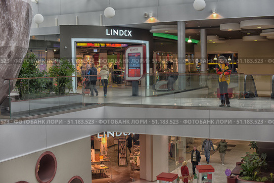 The photo was taken inside a modern shopping center. In the foreground, two levels of the building are visible with spacious corridors decorated with green plants. On the upper floor, there is an interactive navigation screen with a map of the shopping center, and next to it stands a woman in a bright yellow jacket. In the center of the frame is the entrance to the LINDEX store with display cases and bright lighting, and part of the same store is also visible on the lower level. Shoppers walk along the corridors, creating an atmosphere of a lively shopping space. The interior is designed in a modern style with elements of glass partitions and metal structures, which emphasizes the style and spaciousness of the building. (2015 год). Редакционное фото, фотограф Анна Кривицкая / Фотобанк Лори