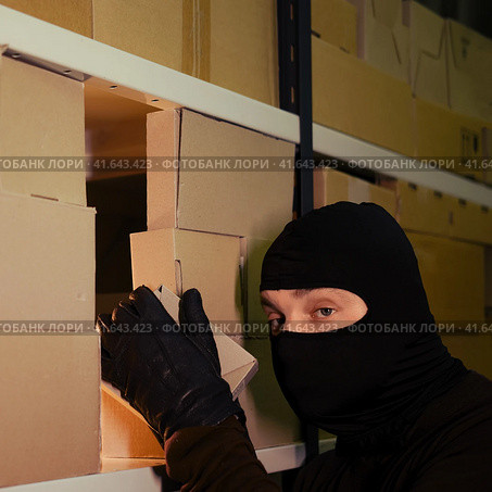 Thief in a black ski mask steals a box with a parcel in a warehouse at night. Concept of security problems in warehouses and stores. Стоковое фото, фотограф Андрей Журавлев / Фотобанк Лори