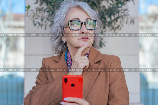 Thoughtful senior woman with smartphone looking away in an urban environment. Стоковое фото, фотограф Raquel Corcoles Mendez / Фотобанк Лори