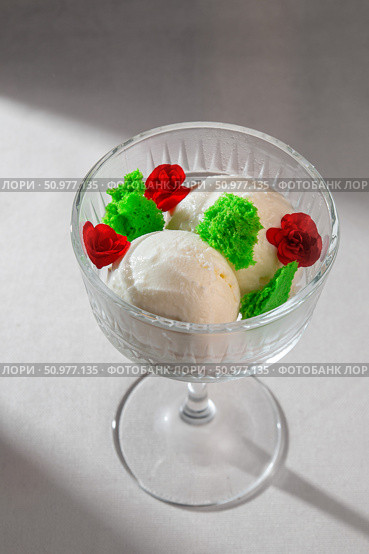 Three creamy scoops of vanilla ice cream beautifully presented in a classic glass dessert bowl, adorned with vibrant red roses and green decorative elements. Стоковое фото, фотограф Aleksandr Gavrilychev / Фотобанк Лори