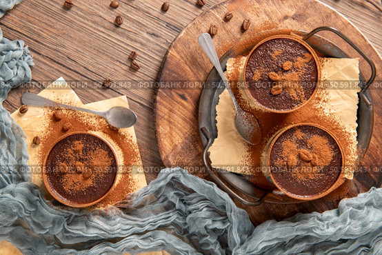 Three delicious chocolate desserts in small bowls, generously dusted with cocoa powder and decorated with chocolate pieces. Set on a rustic wooden tray with spoons and coffee beans, perfect for a delightful treat. Стоковое фото, агентство Ingram Publishing / Фотобанк Лори