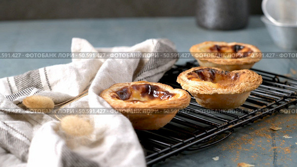 Three golden-brown Portuguese custard tarts, fresh from the oven, cooling on a black wire rack with a white and gray towel on the side. Стоковое фото, агентство Ingram Publishing / Фотобанк Лори