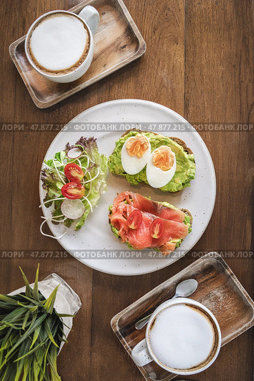 Toasts with avocado, smoked salmon and boiled eggs, salad and 2 cups of cappuccino coffee on a wooden table top view flatlay. Healthy and delicious breakfast for two with coffee or coffee break. Стоковое фото, фотограф Владимир Разгуляев / Фотобанк Лори